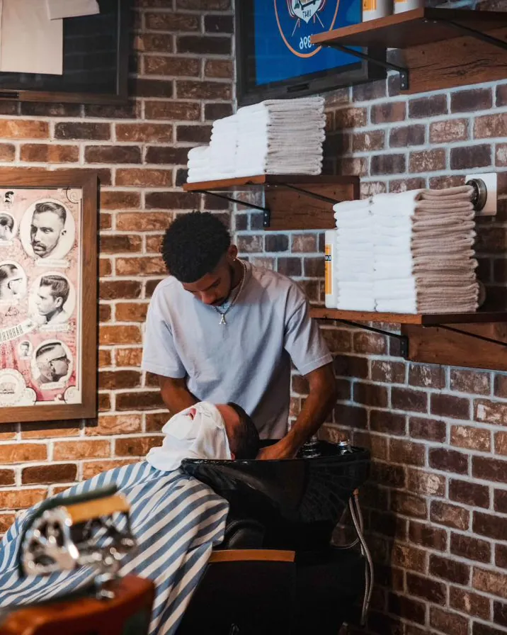 A barber at the wash basin with a client, beneath stacked towels