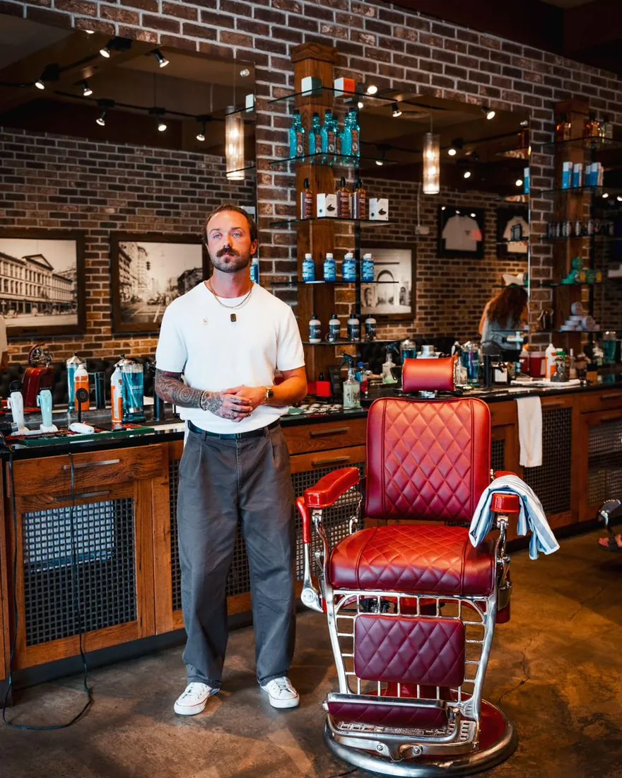 A bearded man standing beside a red barber chair inside City Barbers