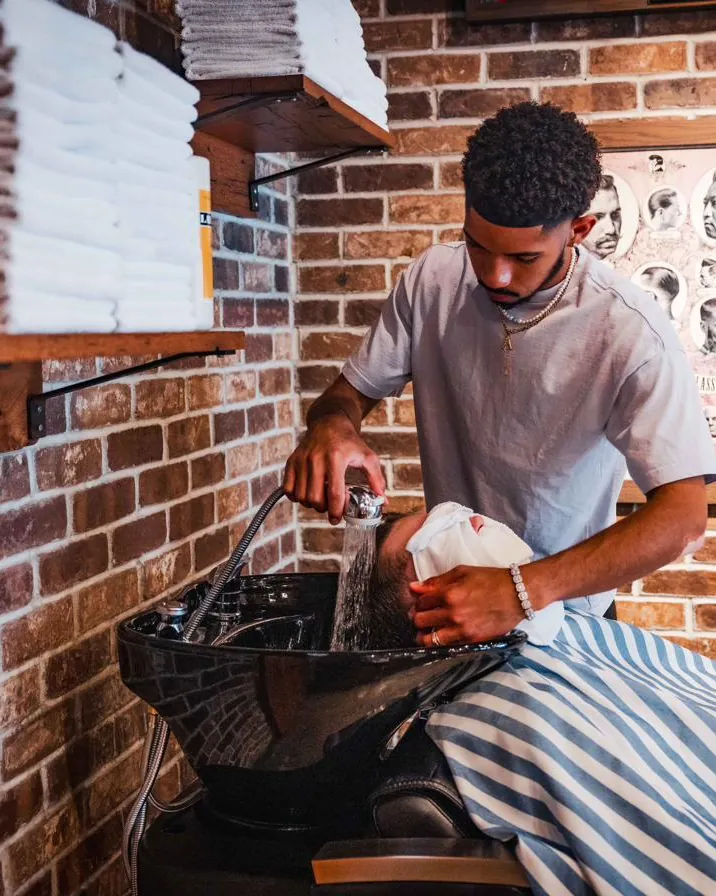 A barber washing a client’s hair at the basin beneath stacked towels