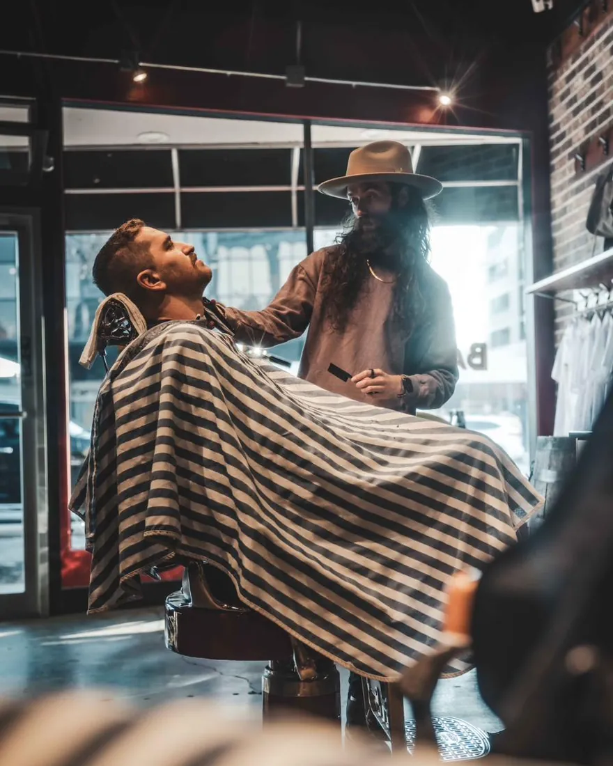 A barber in a wide-brimmed hat working with a bearded client by the front window